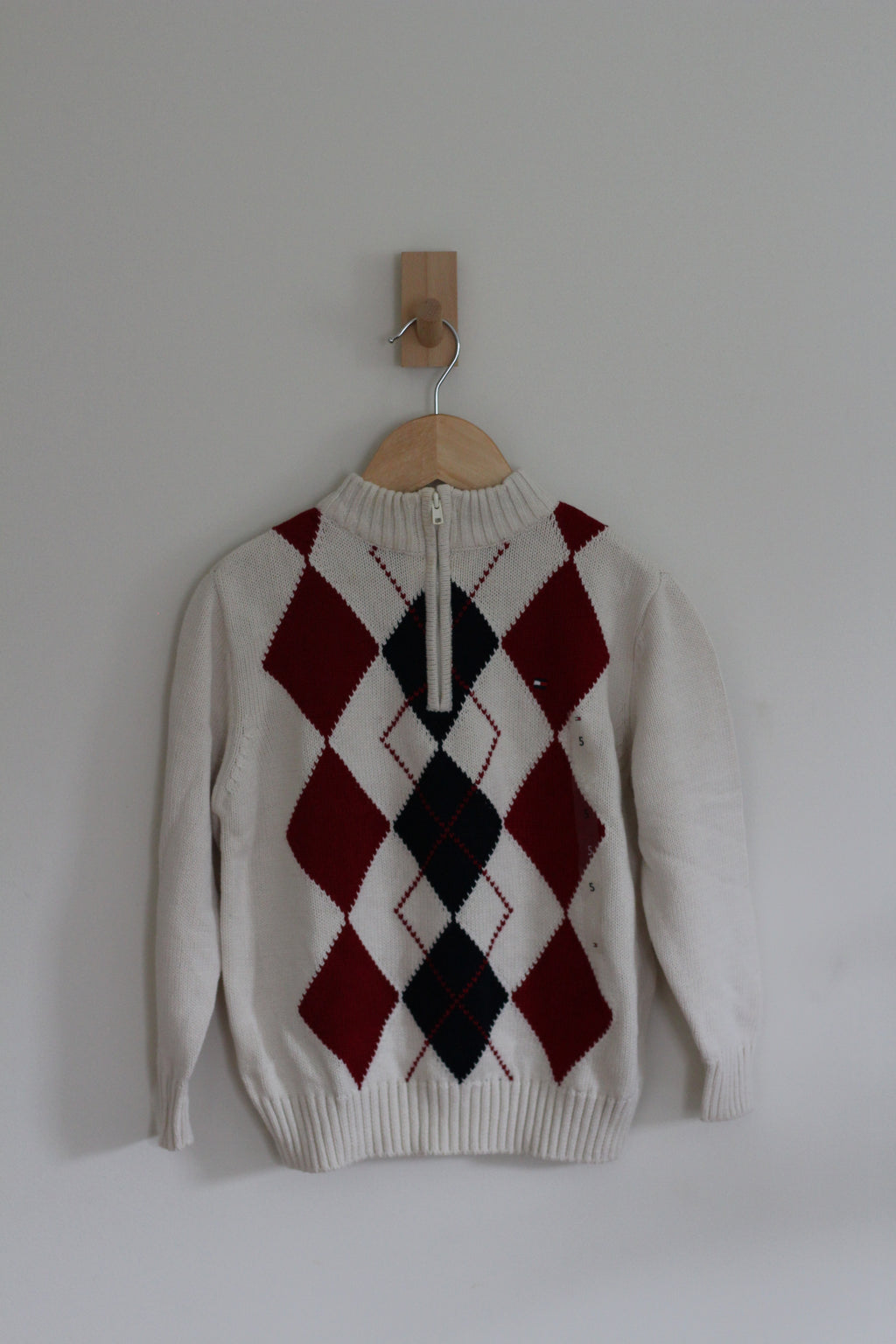 A cream-colored knit sweater with a red and black argyle pattern on the front, hanging on a wooden hanger from a wall hook against a plain light gray wall.
