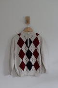 A cream-colored knit sweater with a red and black argyle pattern on the front, hanging on a wooden hanger from a wall hook against a plain light gray wall.