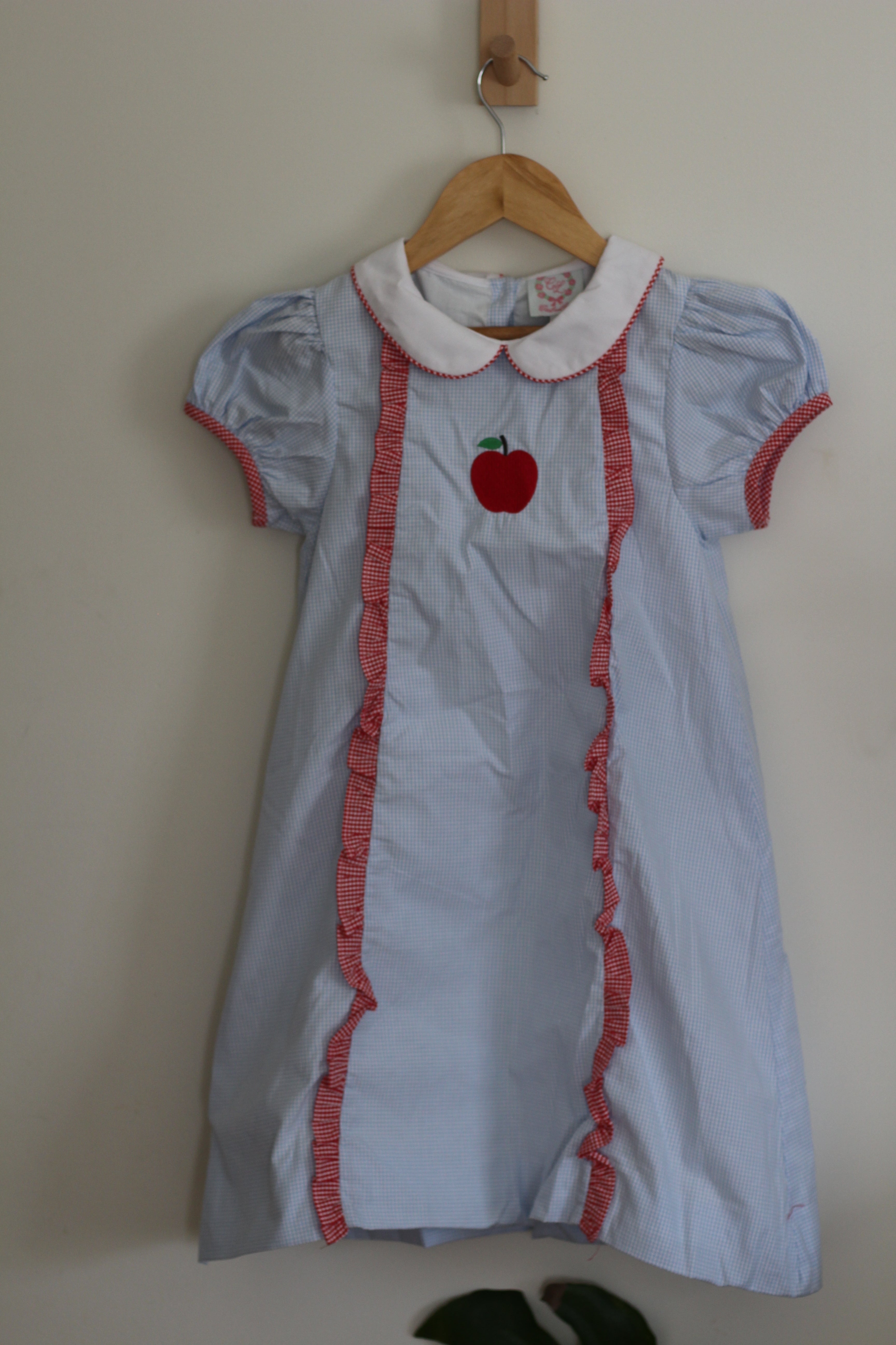 A light blue short-sleeve  Cecil and Lou dress with a white Peter Pan collar, red ruffle trim, and a red apple applique on the chest, hanging on a wooden hanger against a plain beige wall.