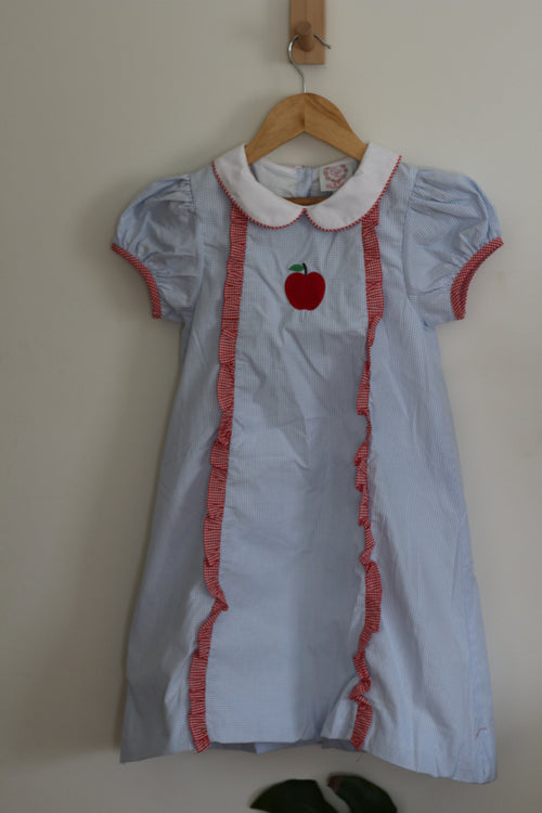 A light blue short-sleeve  Cecil and Lou dress with a white Peter Pan collar, red ruffle trim, and a red apple applique on the chest, hanging on a wooden hanger against a plain beige wall.