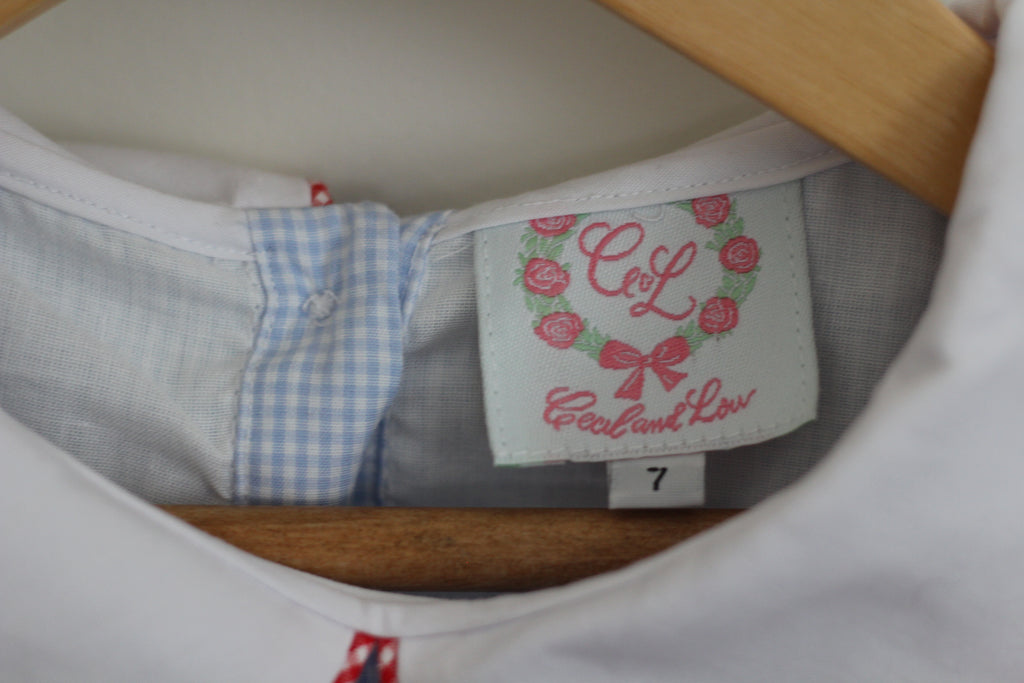 Close-up of the inside neck label of a white shirt on a wooden hanger, showing the brand 'Cecil and Lou with a pink rose wreath logo and a light blue gingham fabric panel.