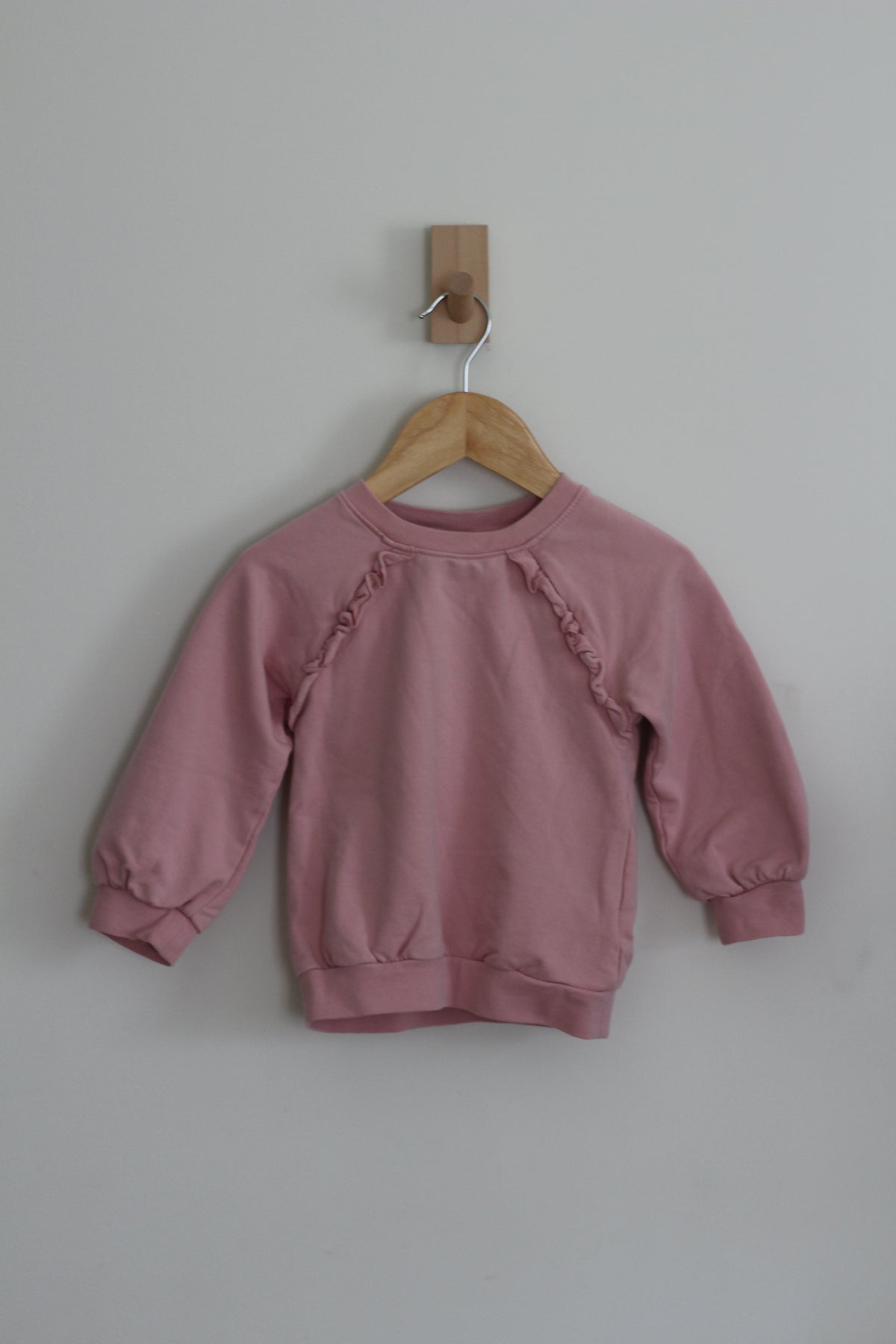 A pink long-sleeve sweatshirt with ruffle details on the shoulders, hanging on a wooden hanger from a wall hook against a light gray wall.