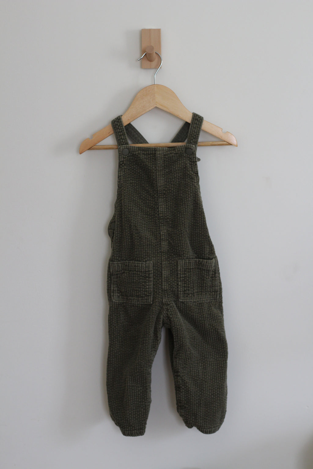 Dark olive green corduroy baby overalls hanging on a wooden hanger against a plain light gray wall.