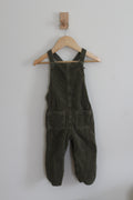 Dark olive green corduroy baby overalls hanging on a wooden hanger against a plain light gray wall.