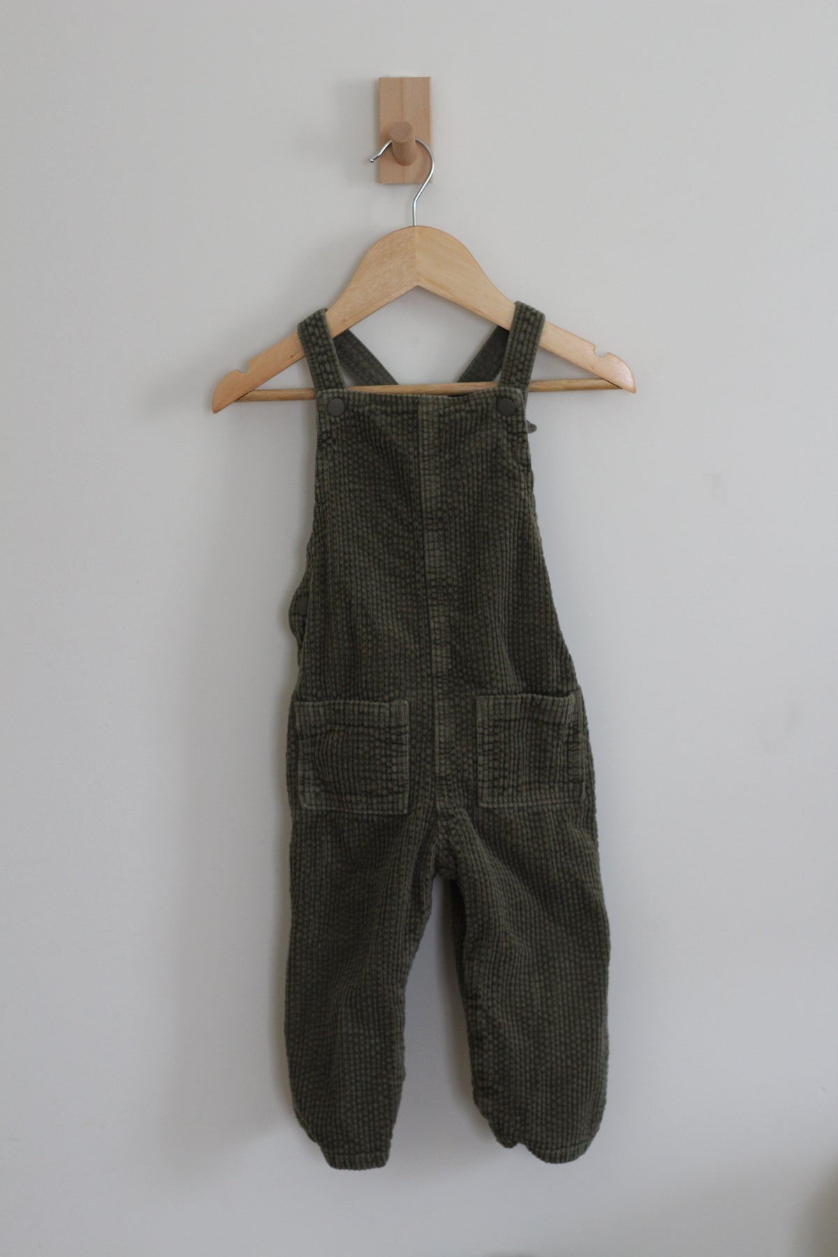 Dark olive green corduroy baby overalls hanging on a wooden hanger against a plain light gray wall.