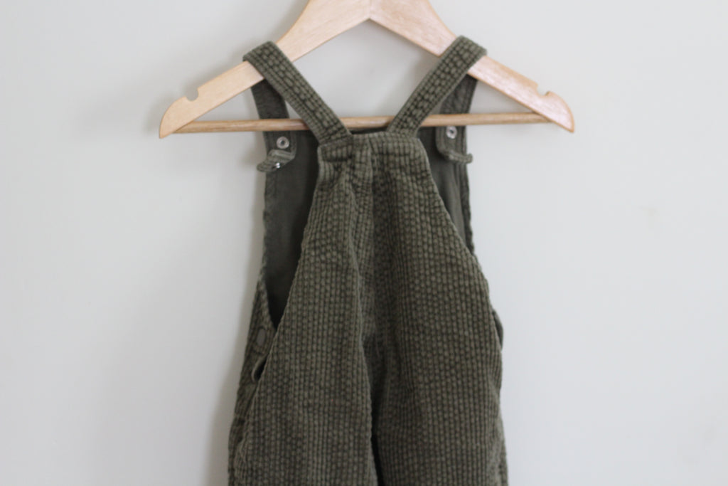 Olive green corduroy bib overalls hanging on a light wood hanger against a plain light gray wall.