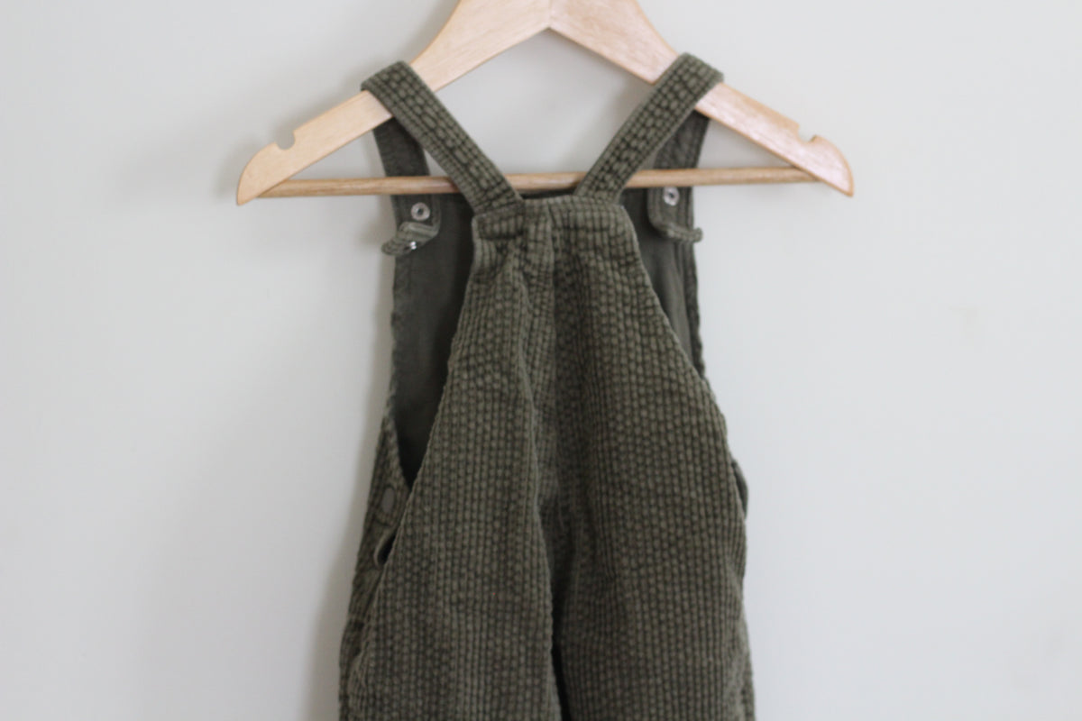 Olive green corduroy bib overalls hanging on a light wood hanger against a plain light gray wall.