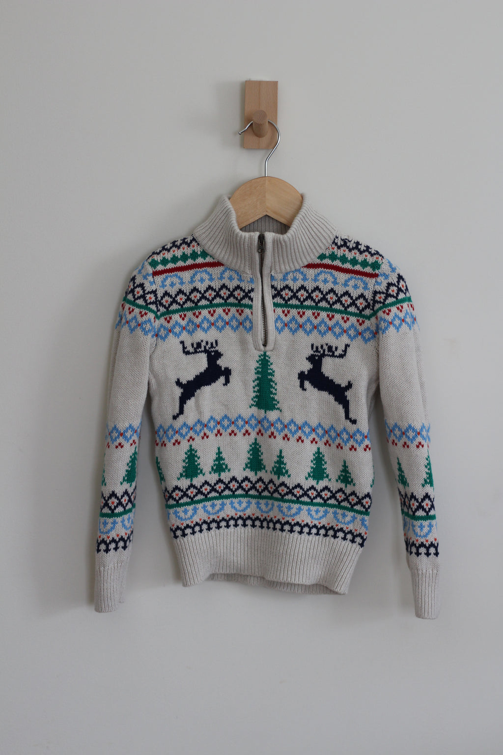A cream-colored knit sweater with a half-zip neckline, featuring a festive Fair Isle pattern with blue, green, and red accents, and two black reindeer motifs on the chest. The sweater is hanging on a wooden hanger from a wooden peg against a plain light gray wall.