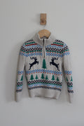 A cream-colored knit sweater with a half-zip neckline, featuring a festive Fair Isle pattern with blue, green, and red accents, and two black reindeer motifs on the chest. The sweater is hanging on a wooden hanger from a wooden peg against a plain light gray wall.