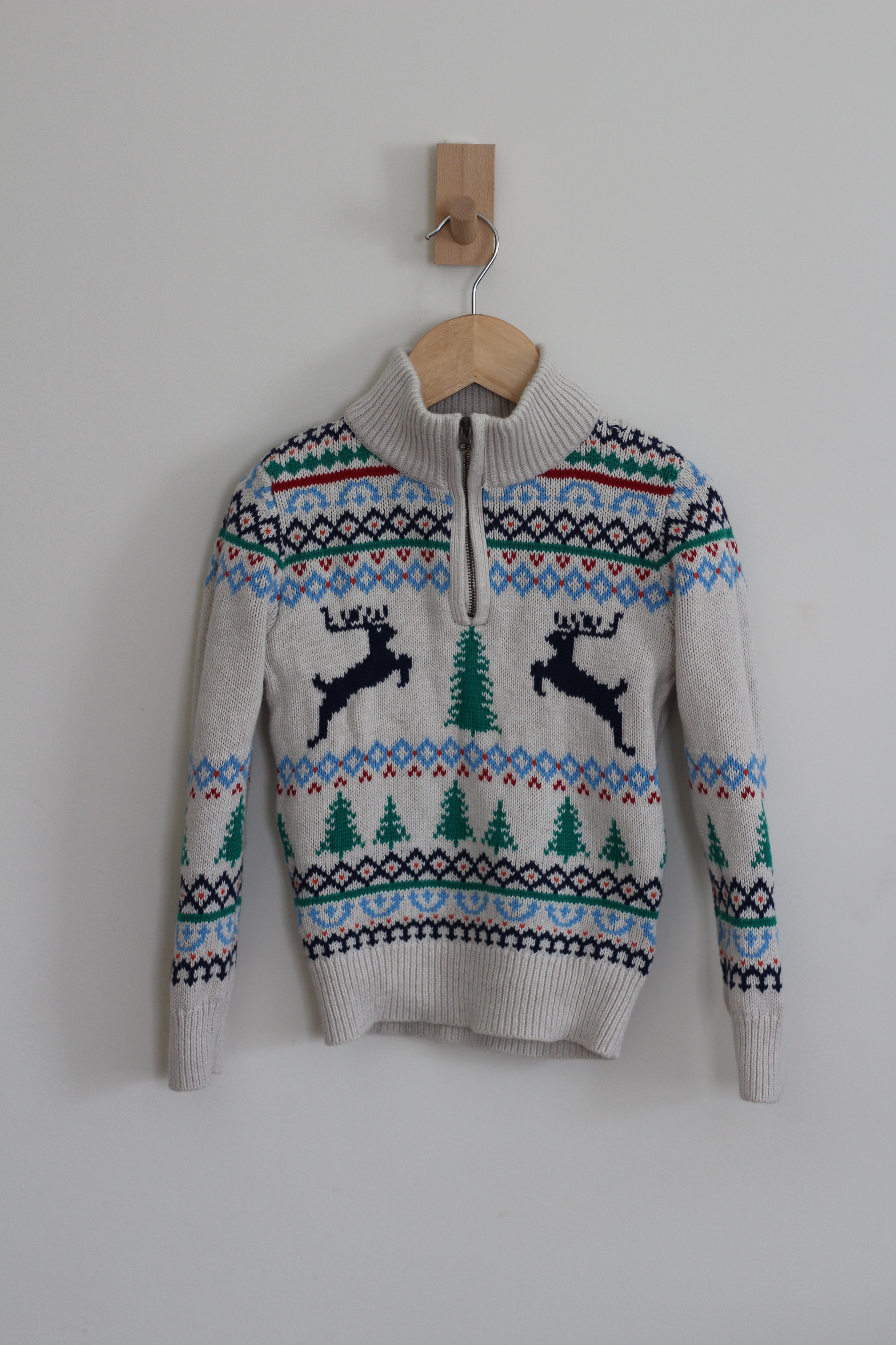 A cream-colored knit sweater with a half-zip neckline, featuring a festive Fair Isle pattern with blue, green, and red accents, and two black reindeer motifs on the chest. The sweater is hanging on a wooden hanger from a wooden peg against a plain light gray wall.