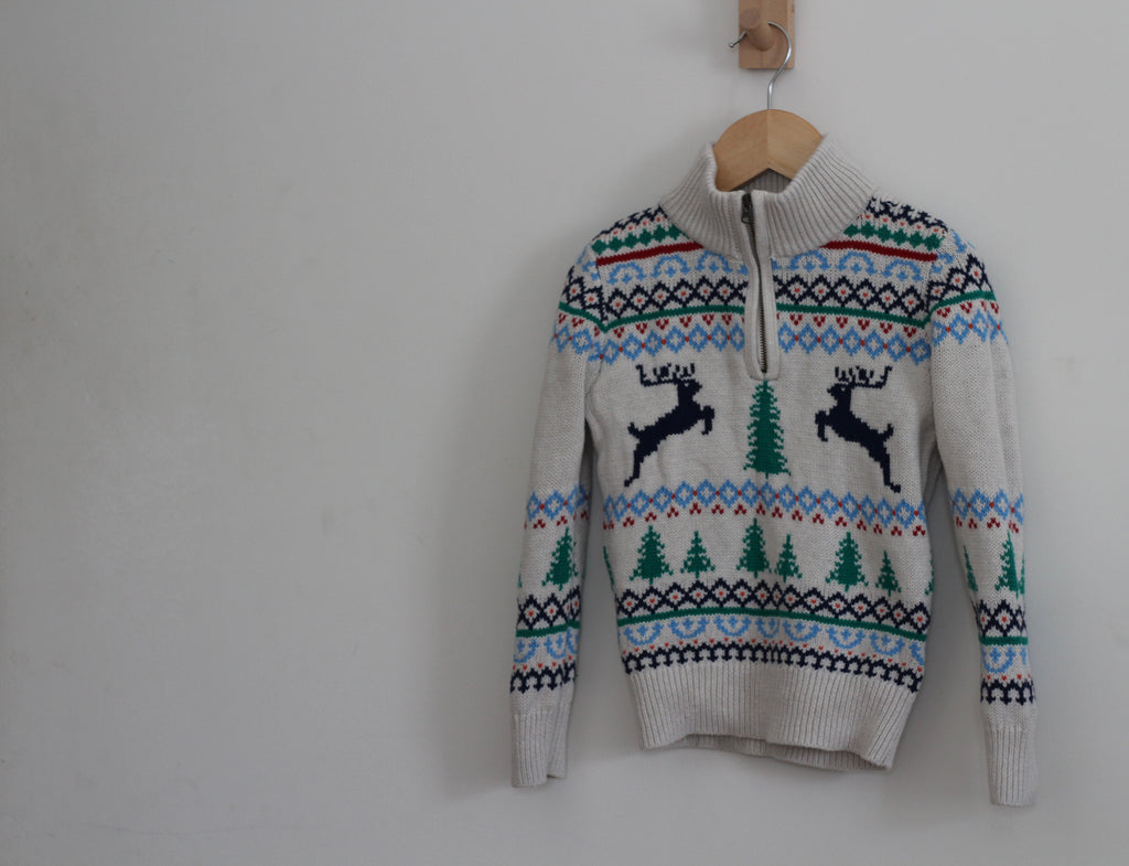 A knit half-zip sweater with a Christmas fair isle pattern featuring two black leaping reindeer and green pine trees, hanging on a wooden hanger against a plain light gray wall.