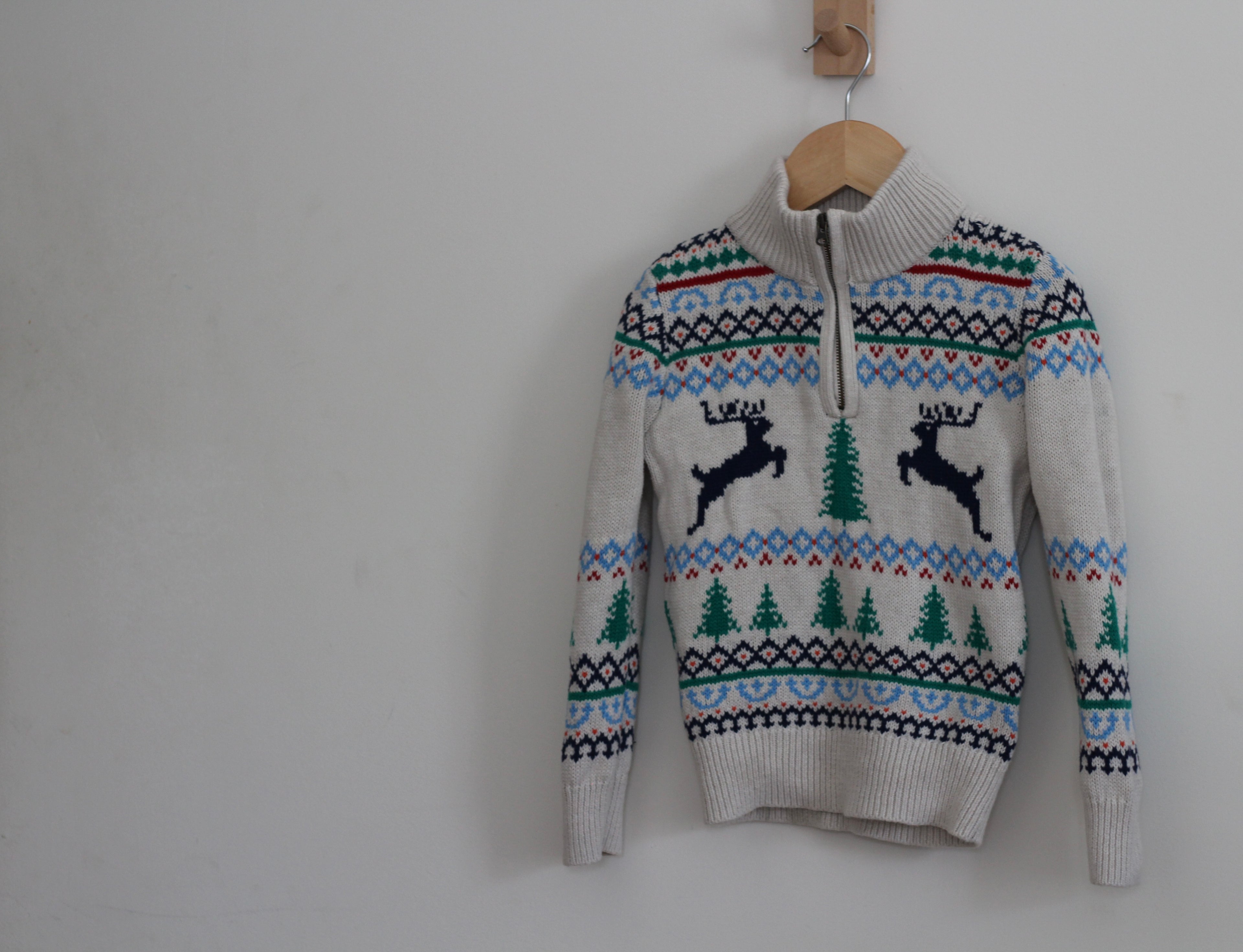 A knit half-zip sweater with a Christmas fair isle pattern featuring two black leaping reindeer and green pine trees, hanging on a wooden hanger against a plain light gray wall.