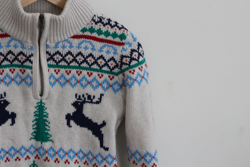 A close-up of a knit half-zip sweater with a cream base and a colorful Nordic holiday pattern featuring blue, green, and red geometric motifs, along with dark blue reindeer silhouettes and a green Christmas tree.