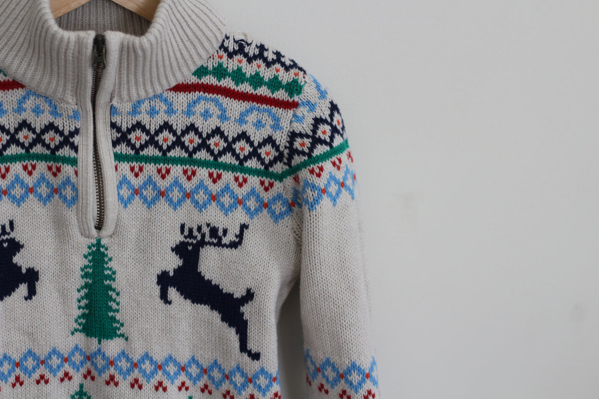 A close-up of a knit half-zip sweater with a cream base and a colorful Nordic holiday pattern featuring blue, green, and red geometric motifs, along with dark blue reindeer silhouettes and a green Christmas tree.