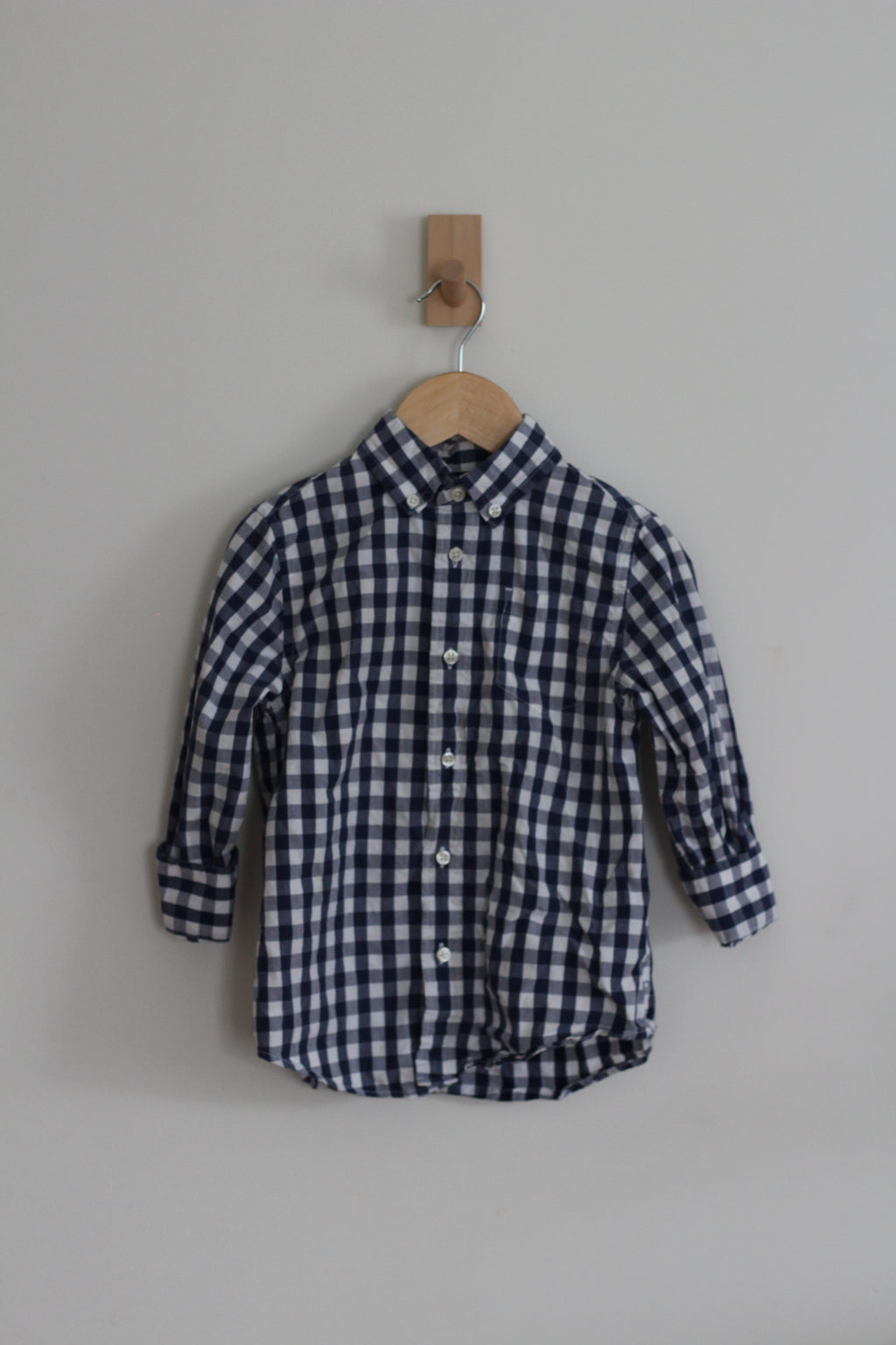 A long-sleeve button-up shirt with a navy and white gingham check pattern, hanging on a wooden hanger from a small wooden wall hook against a plain light gray wall.