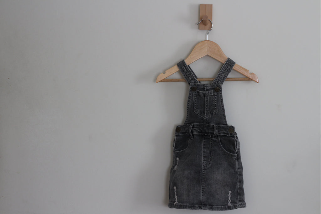 A dark gray washed denim overall dress with front pockets, hanging on a wooden hanger from a wall hook against a plain light gray wall.