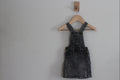 A dark gray washed denim overall dress with front pockets, hanging on a wooden hanger from a wall hook against a plain light gray wall.