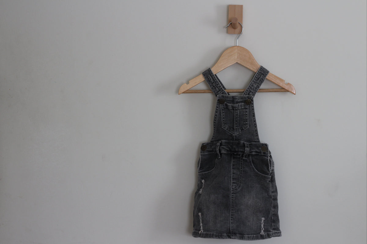 A dark gray washed denim overall dress with front pockets, hanging on a wooden hanger from a wall hook against a plain light gray wall.