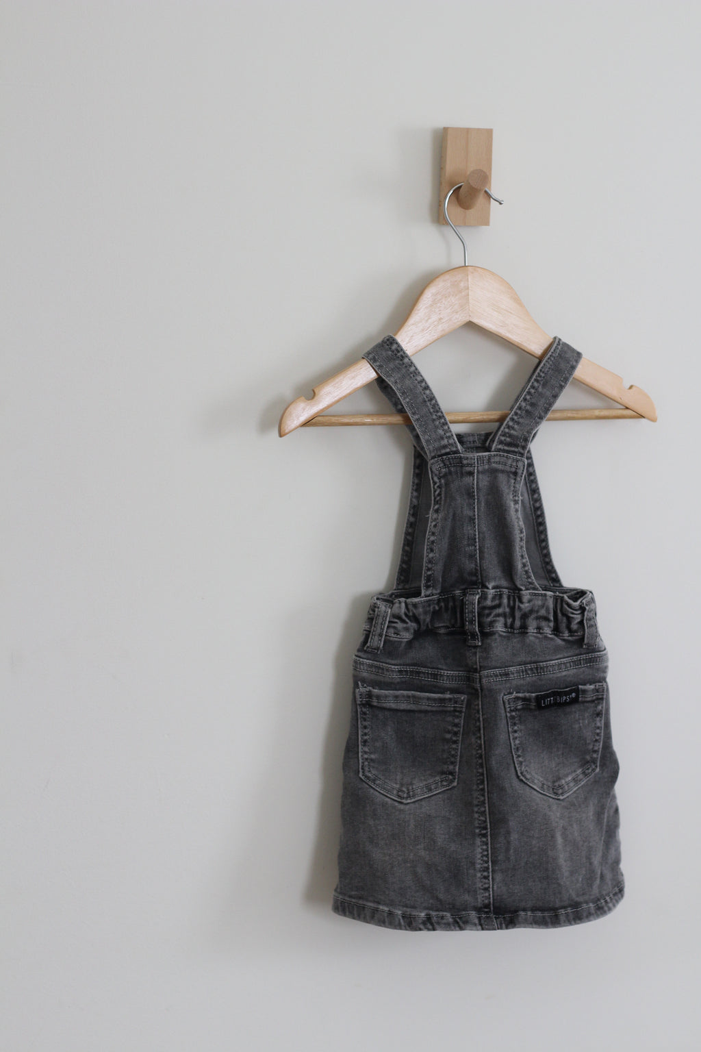A baby girl's dark gray washed denim overall dress hanging on a wooden hanger from a wall hook against a light gray wall.