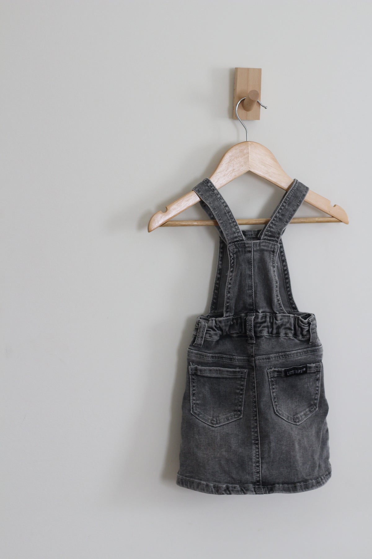 A baby girl's dark gray washed denim overall dress hanging on a wooden hanger from a wall hook against a light gray wall.
