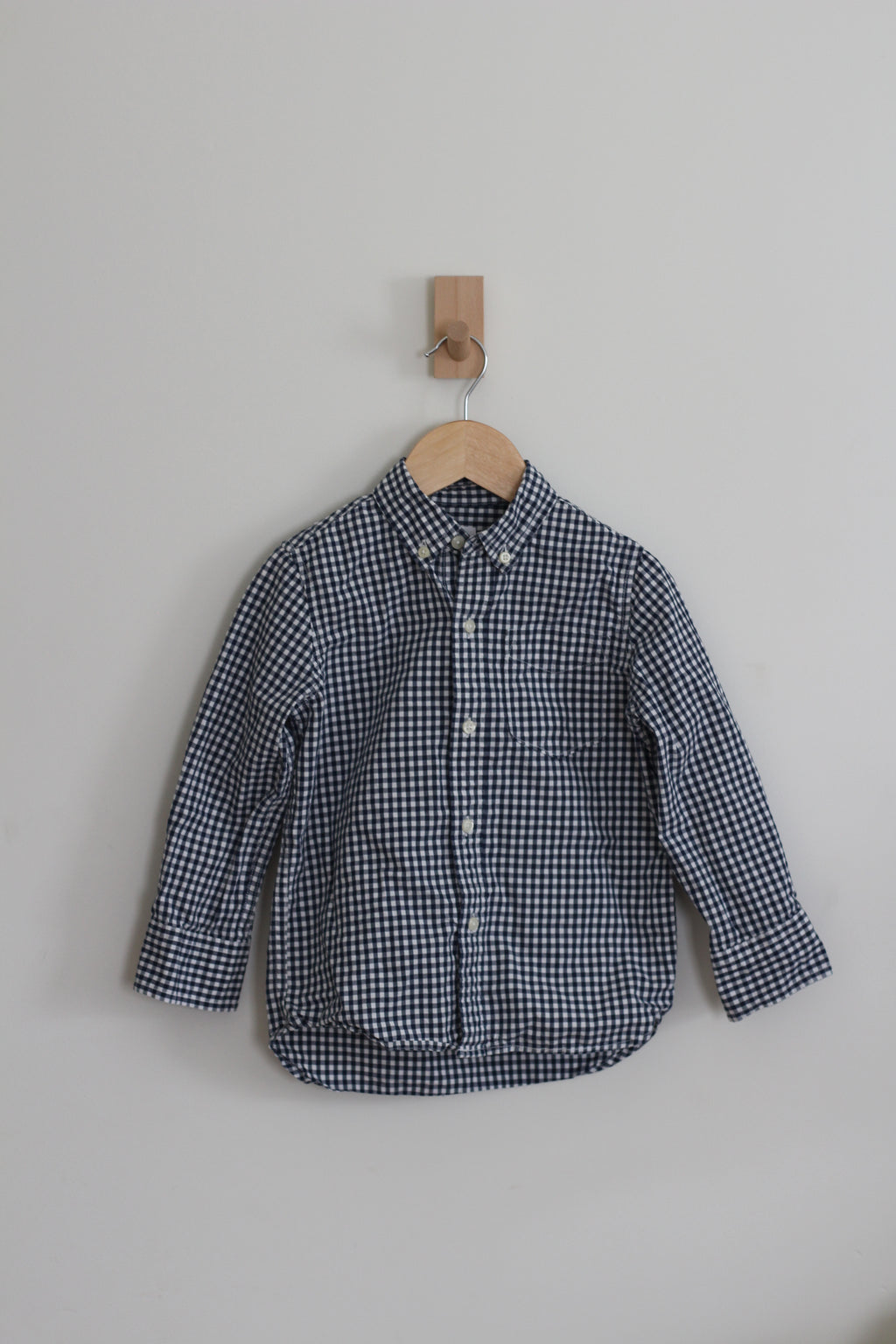 A long-sleeve button-up shirt with a small navy and white gingham check pattern, hanging on a wooden hanger from a wall hook against a plain light gray wall.