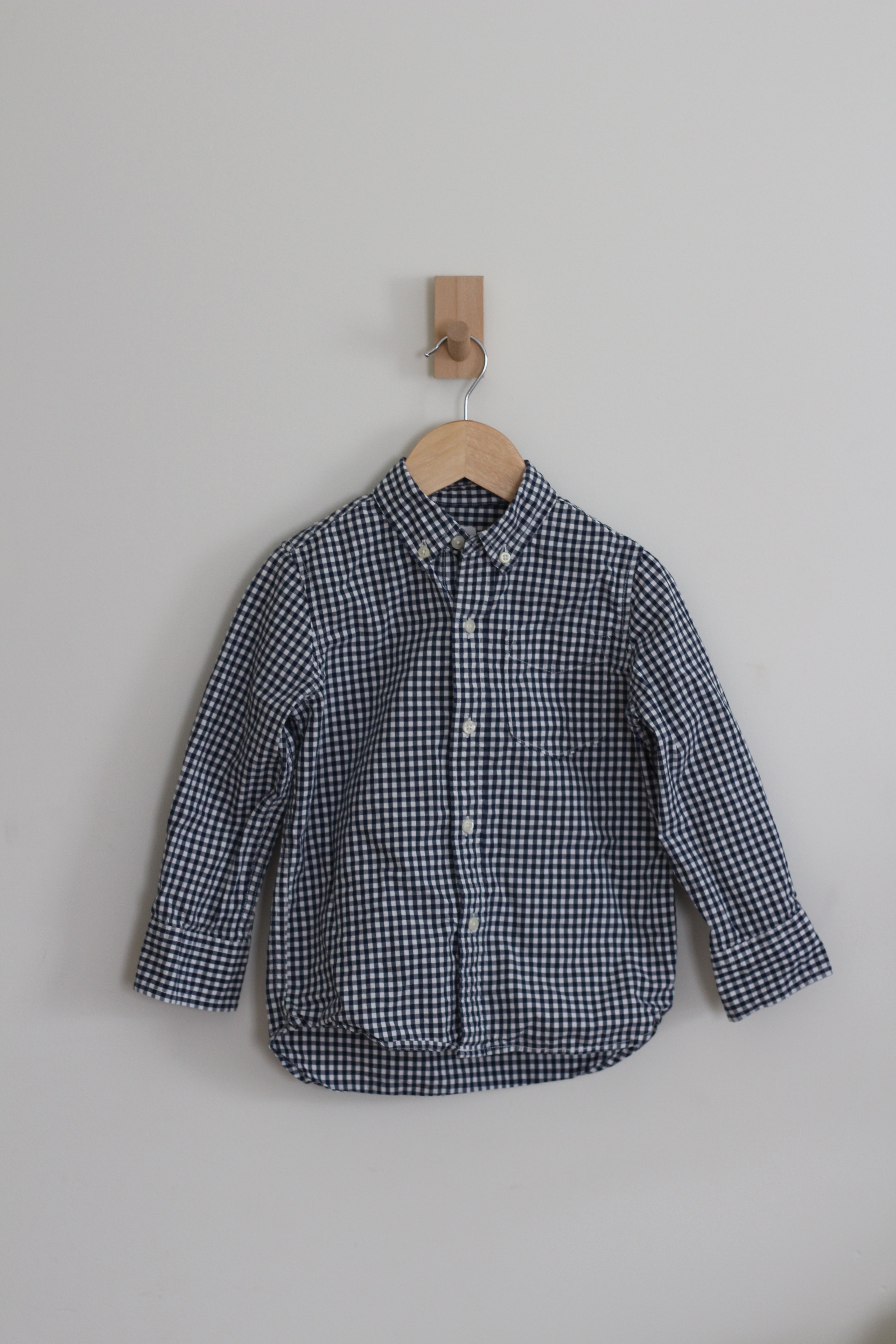 A long-sleeve button-up shirt with a small navy and white gingham check pattern, hanging on a wooden hanger from a wall hook against a plain light gray wall.
