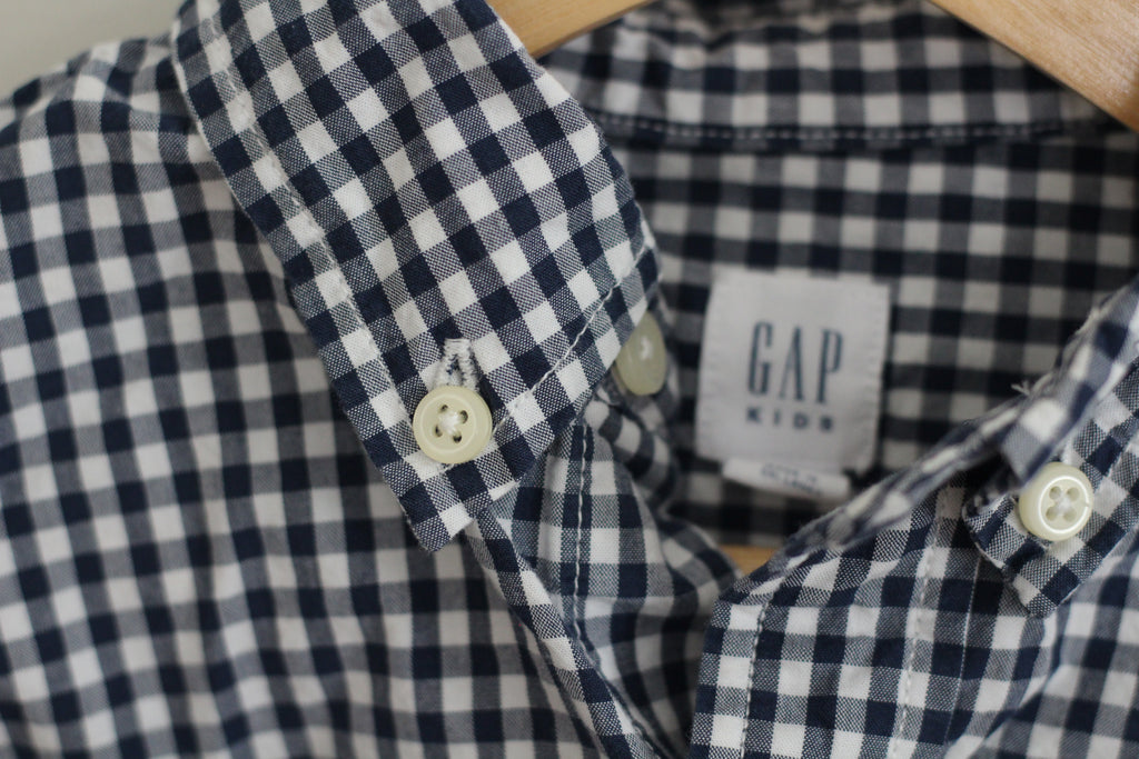 Close-up of a navy and white gingham children's button-down shirt on a wooden hanger, showing the collar, buttons, and a visible GAP Kids brand label.