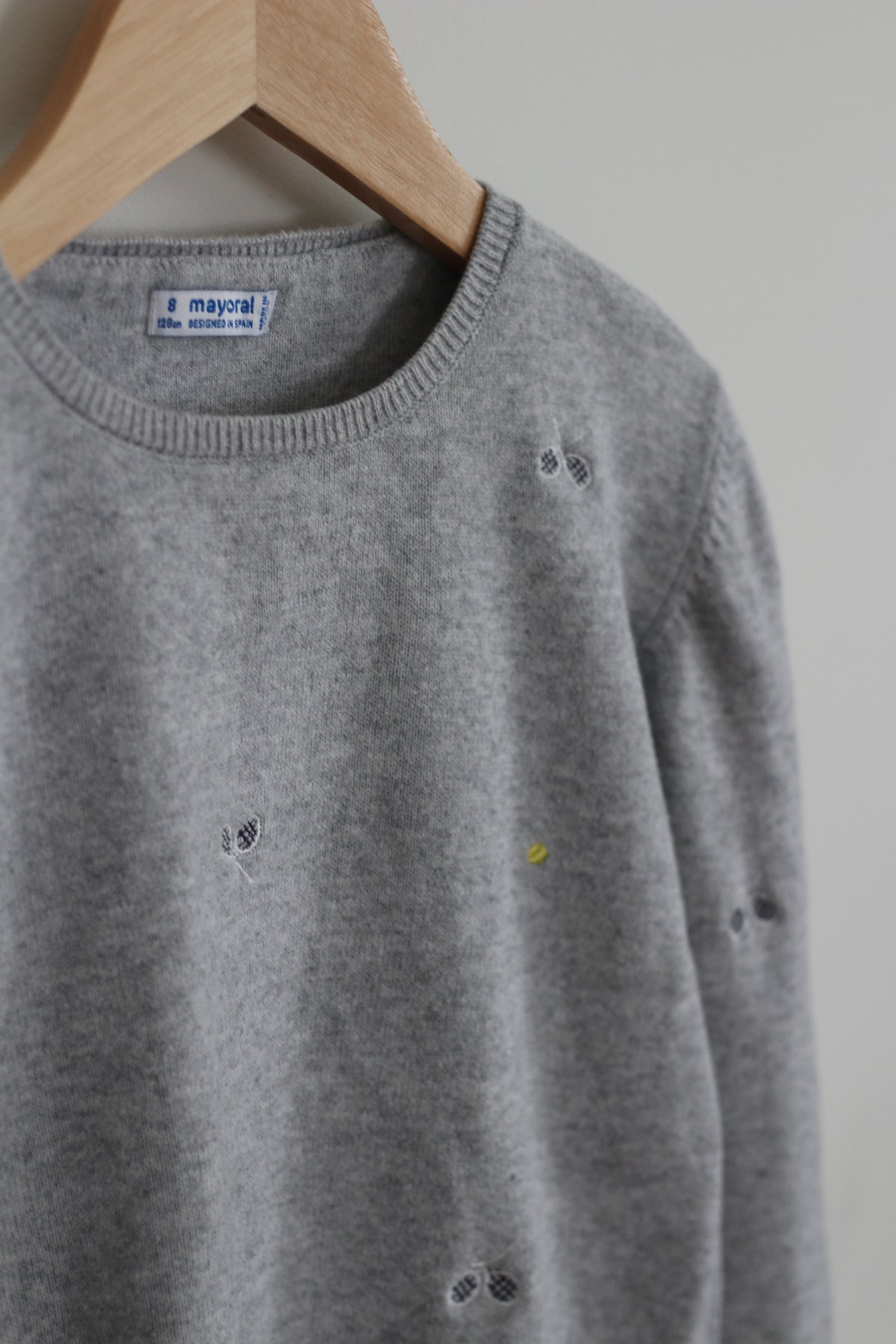 A close-up of a gray knit sweater hanging on a wooden hanger against a light gray wall. The sweater has small scattered dark and yellow embroidered details, and a visible neck label reads 'i mayoral'.