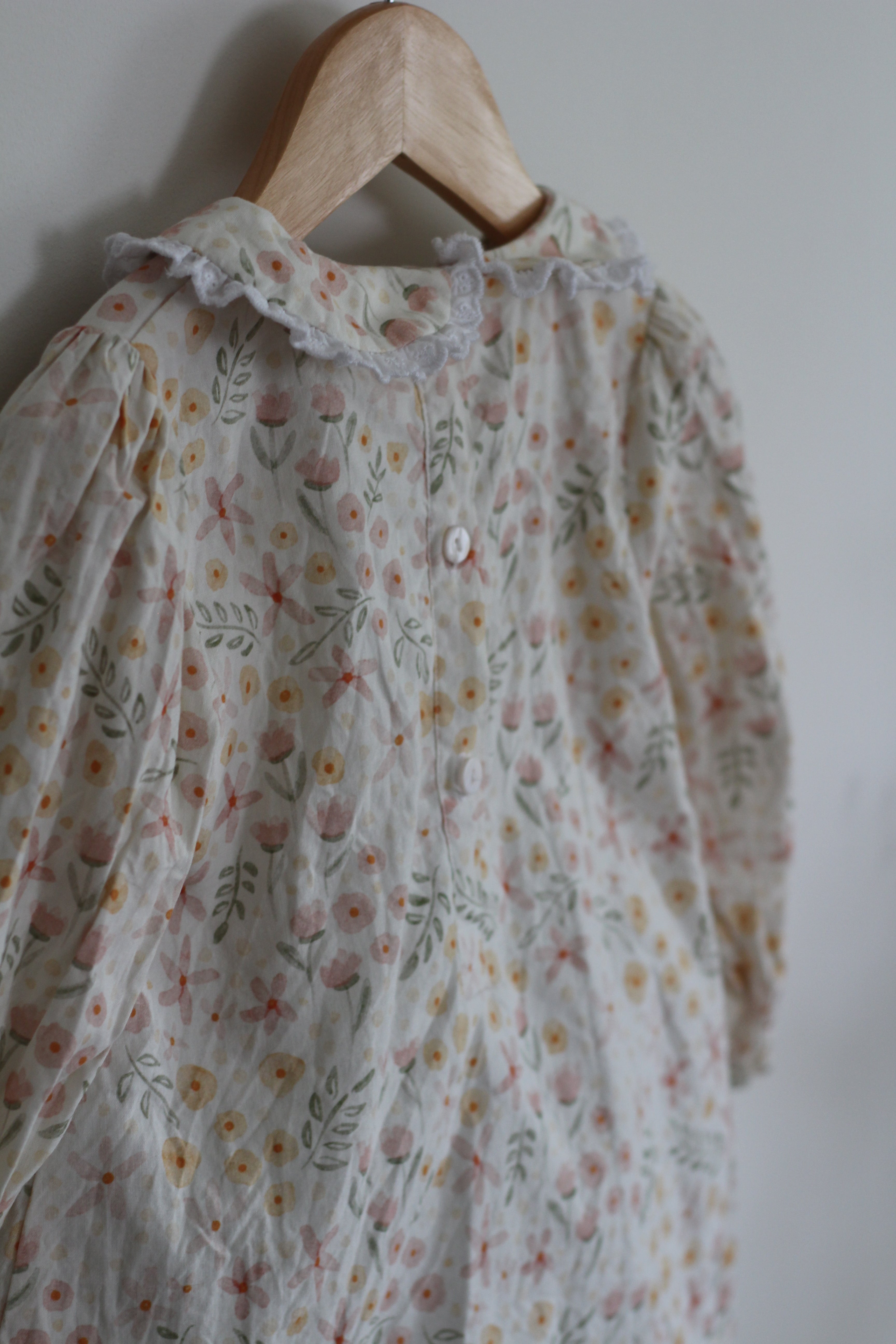 A long-sleeve baby dress with a delicate floral print, hanging on a wooden hanger against a plain light gray wall.