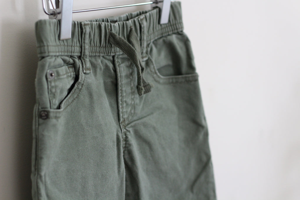 Olive green drawstring shorts hanging from a metal hanger against a plain white wall.