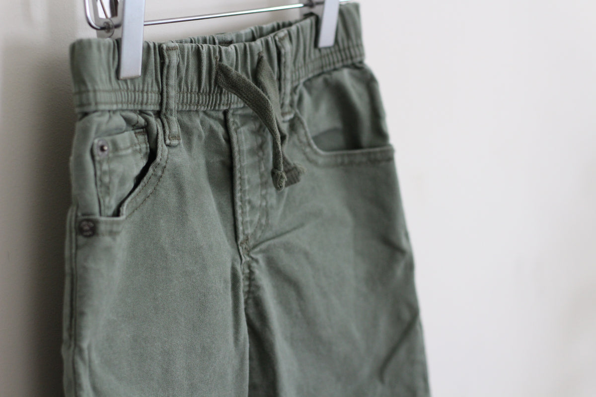 Olive green drawstring shorts hanging from a metal hanger against a plain white wall.