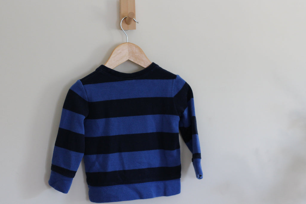 A blue and black striped long-sleeve knit top hanging on a wooden hanger from a wall peg against a plain light wall.