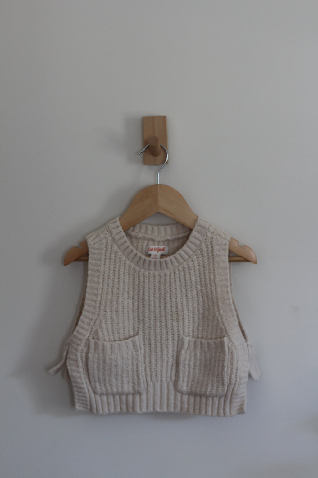 A cream-colored ribbed knit vest with two front patch pockets, hanging on a wooden hanger from a wall peg against a plain light gray wall.
