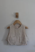A cream-colored ribbed knit vest with two front patch pockets, hanging on a wooden hanger from a wall peg against a plain light gray wall.
