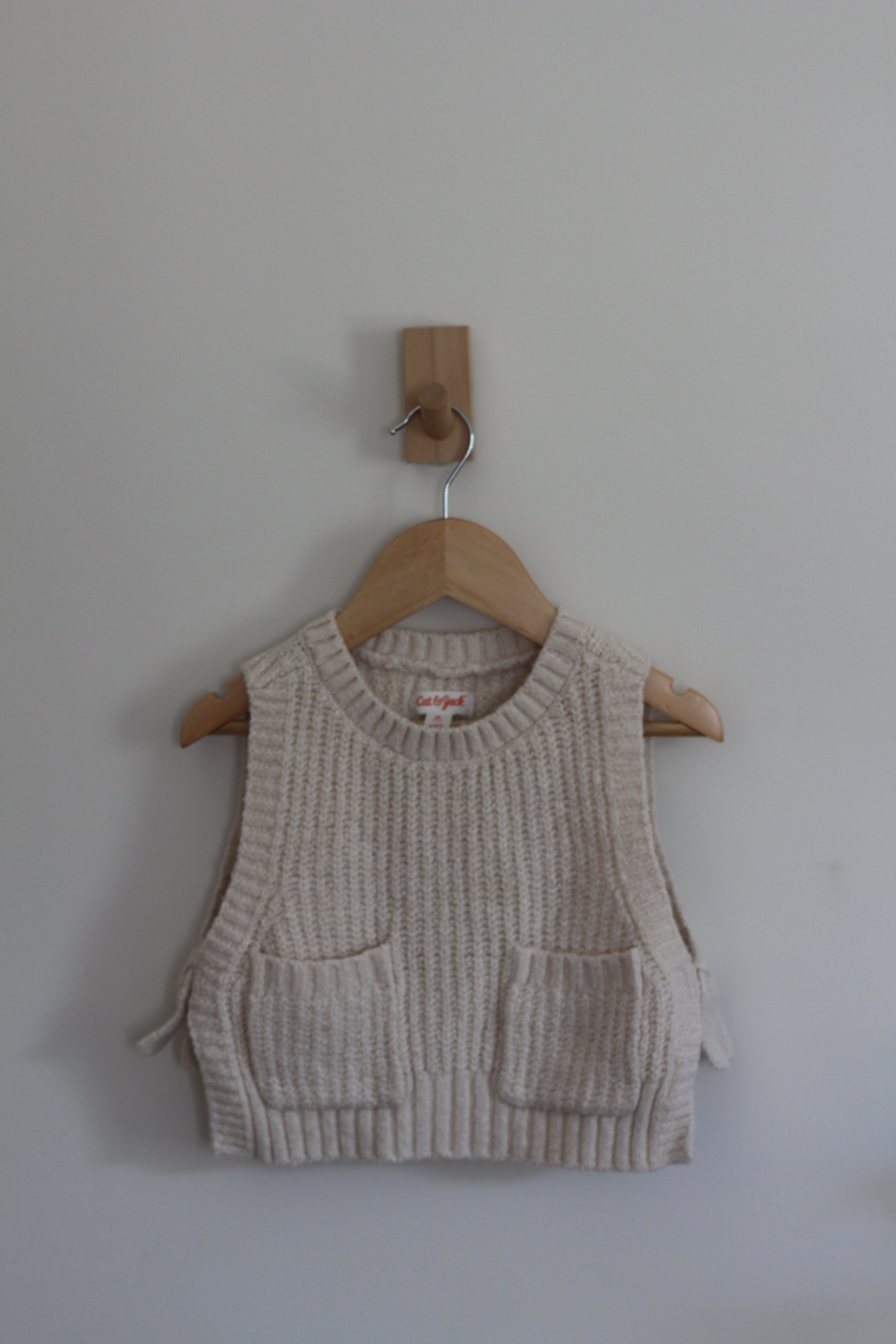A cream-colored ribbed knit vest with two front patch pockets, hanging on a wooden hanger from a wall peg against a plain light gray wall.