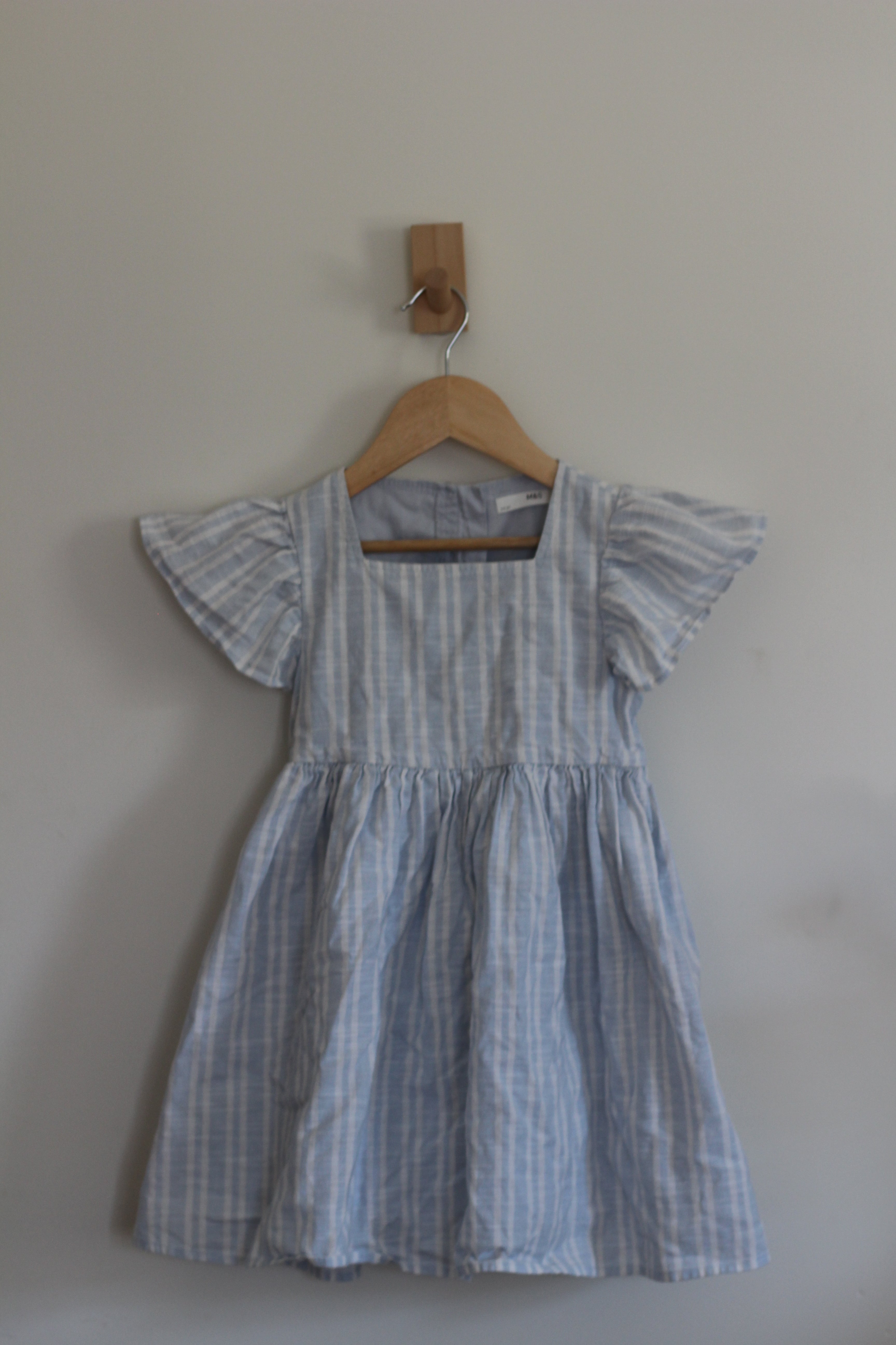 A light blue and white vertically striped baby dress with ruffled flutter sleeves, hanging on a wooden hanger from a wooden peg against a plain light gray wall.