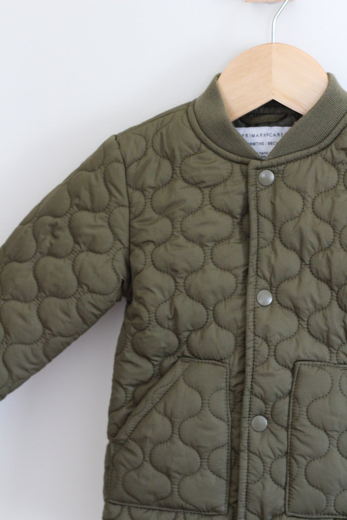 An olive green quilted jacket with a scalloped pattern, featuring a button-up front and a ribbed collar, hanging on a wooden hanger against a plain white wall.