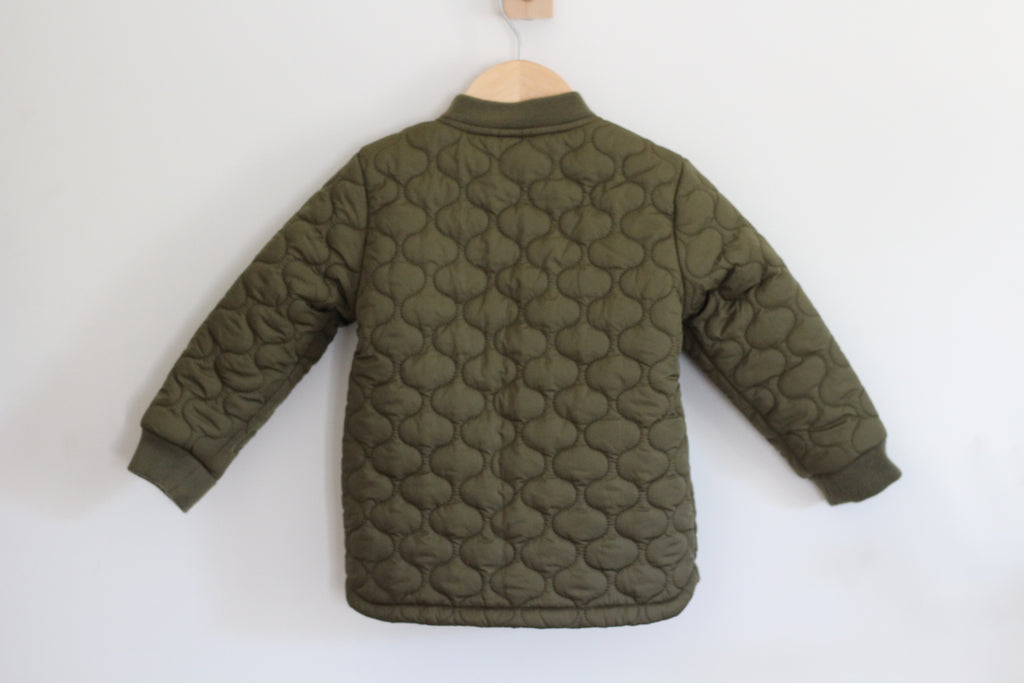 An olive green quilted jacket with a scallop pattern, hanging on a wooden hanger against a plain white wall.