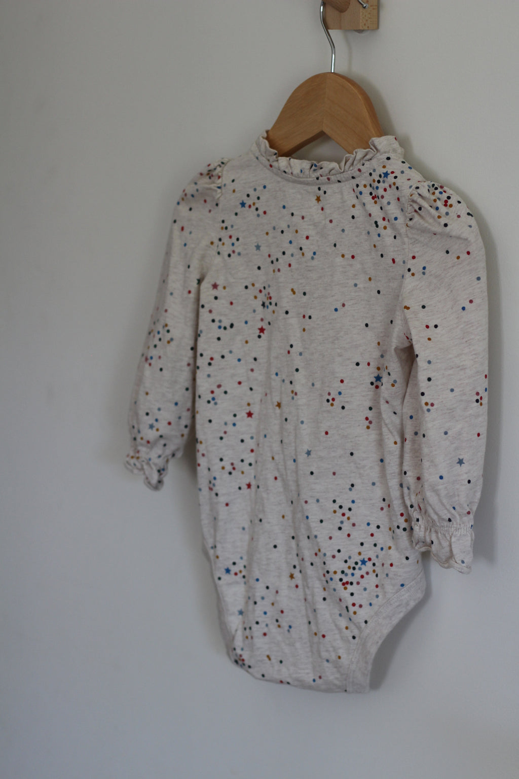 A baby long-sleeve bodysuit in a light heathered cream color, decorated with a scattered pattern of small multicolored dots, hanging on a wooden hanger against a plain light gray wall.