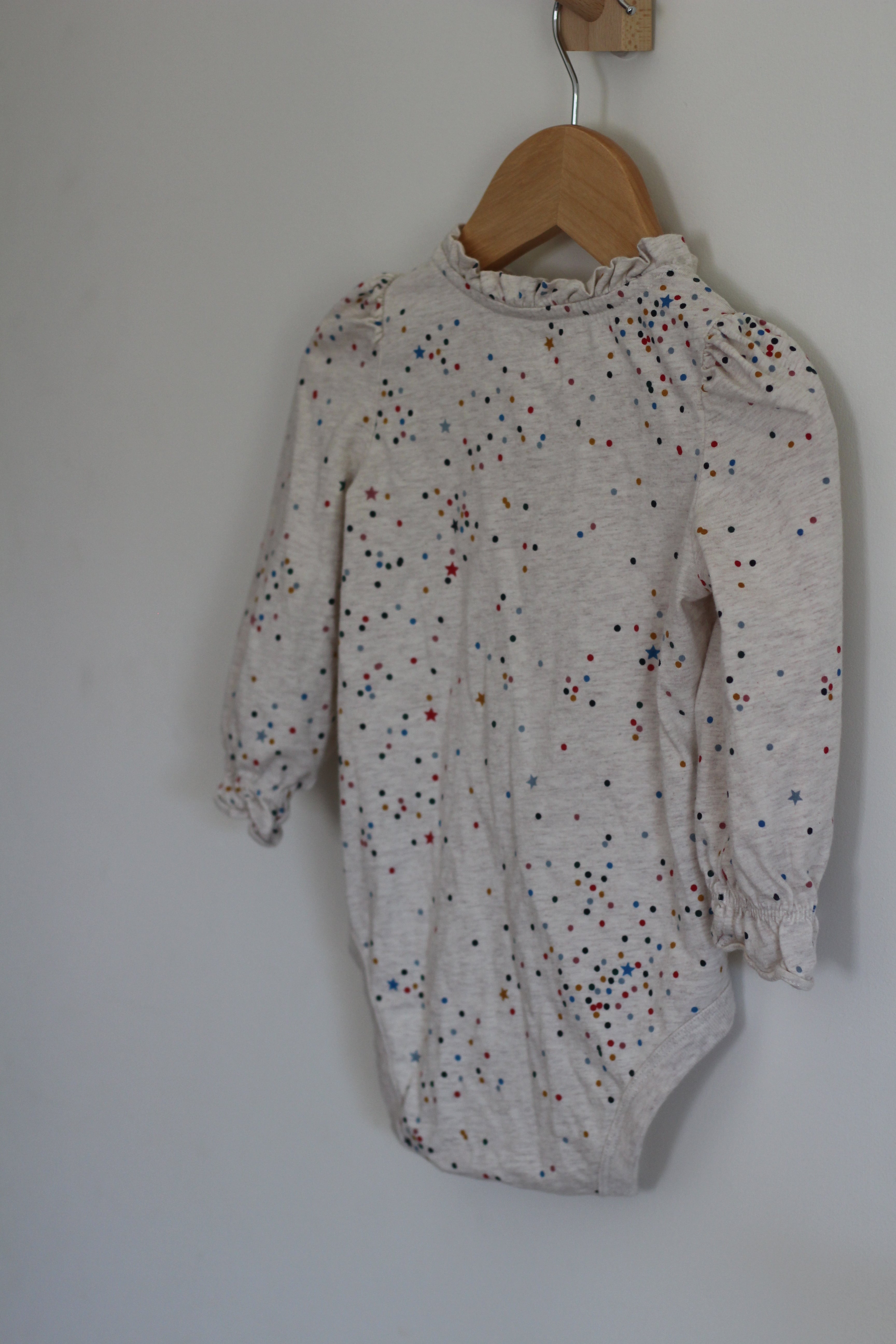 A baby long-sleeve bodysuit in a light heathered cream color, decorated with a scattered pattern of small multicolored dots, hanging on a wooden hanger against a plain light gray wall.