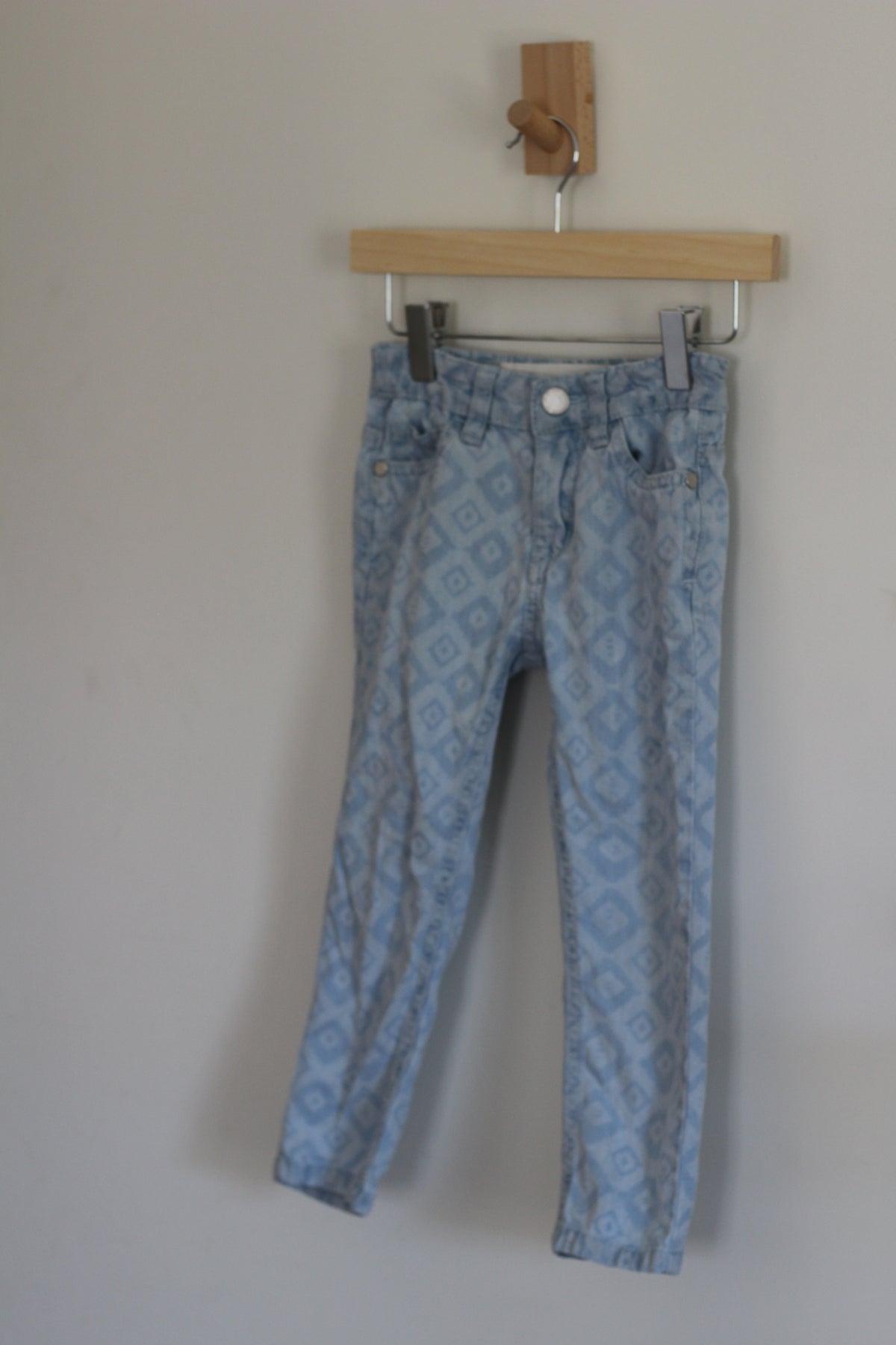 Light blue patterned denim jeans hanging from a wooden hanger on a wall.