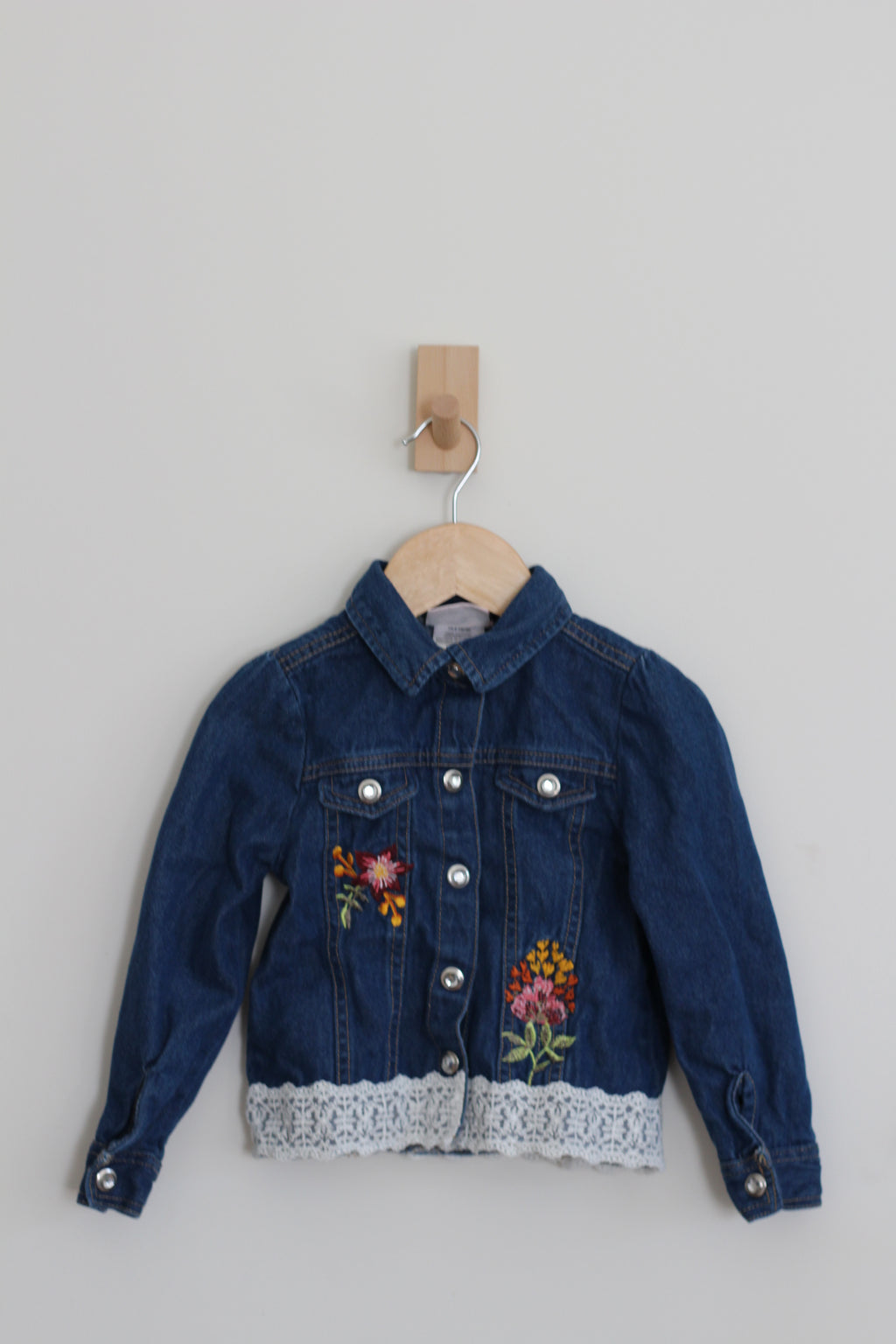 A dark blue denim jacket for kids, featuring colorful floral embroidery on the front, a white lace hem, and silver snap buttons, hanging on a wooden hanger against a light gray wall.