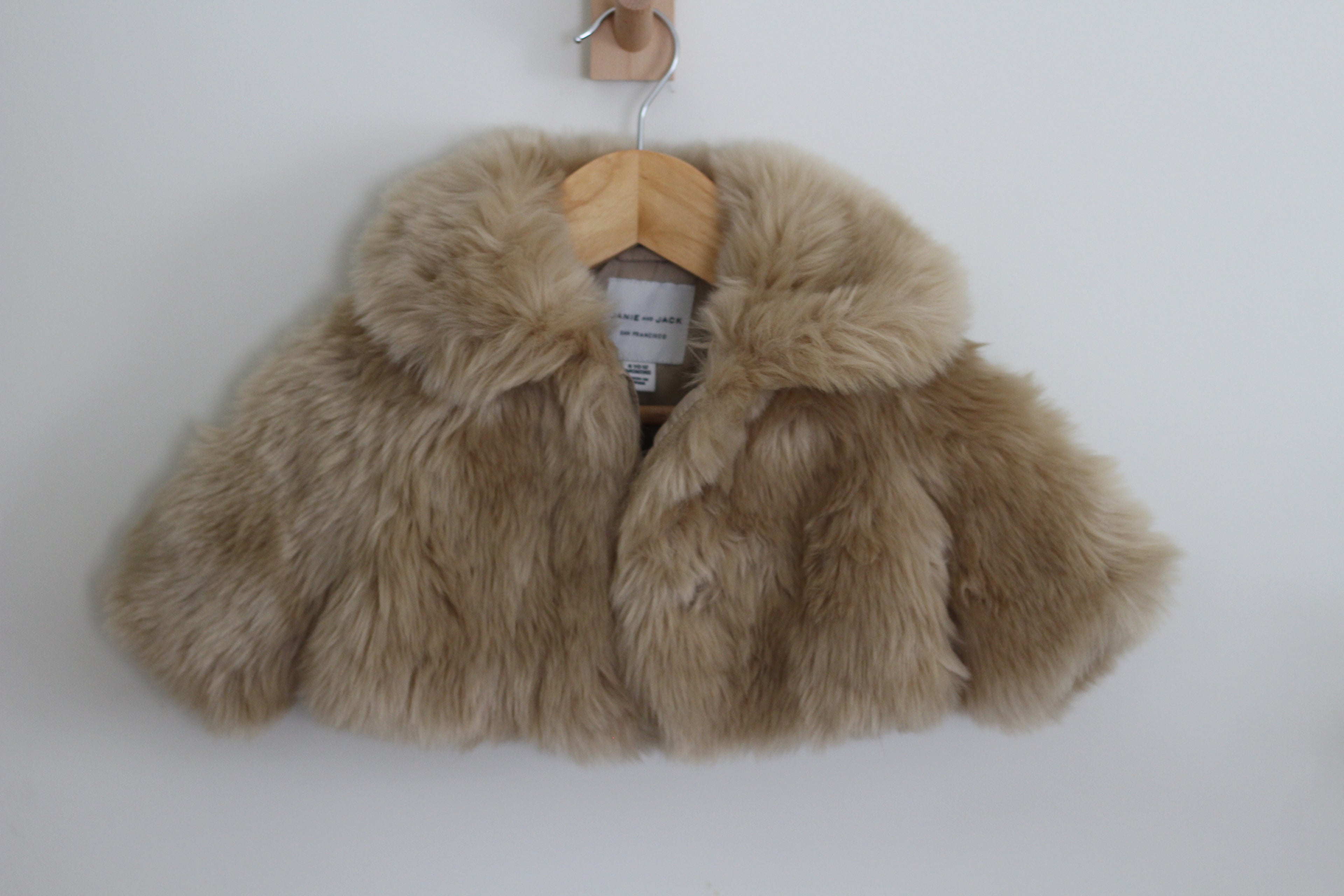 A cropped faux fur jacket by Janie and Jack in a light tan/beige color, hanging on a wooden hanger against a plain white wall.