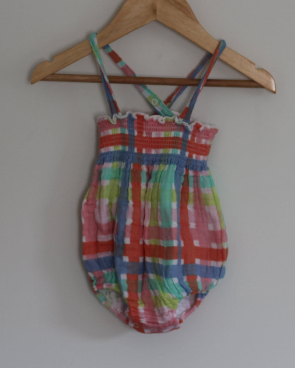 A colorful plaid baby romper with thin shoulder straps, hanging on a wooden hanger against a plain light gray wall.