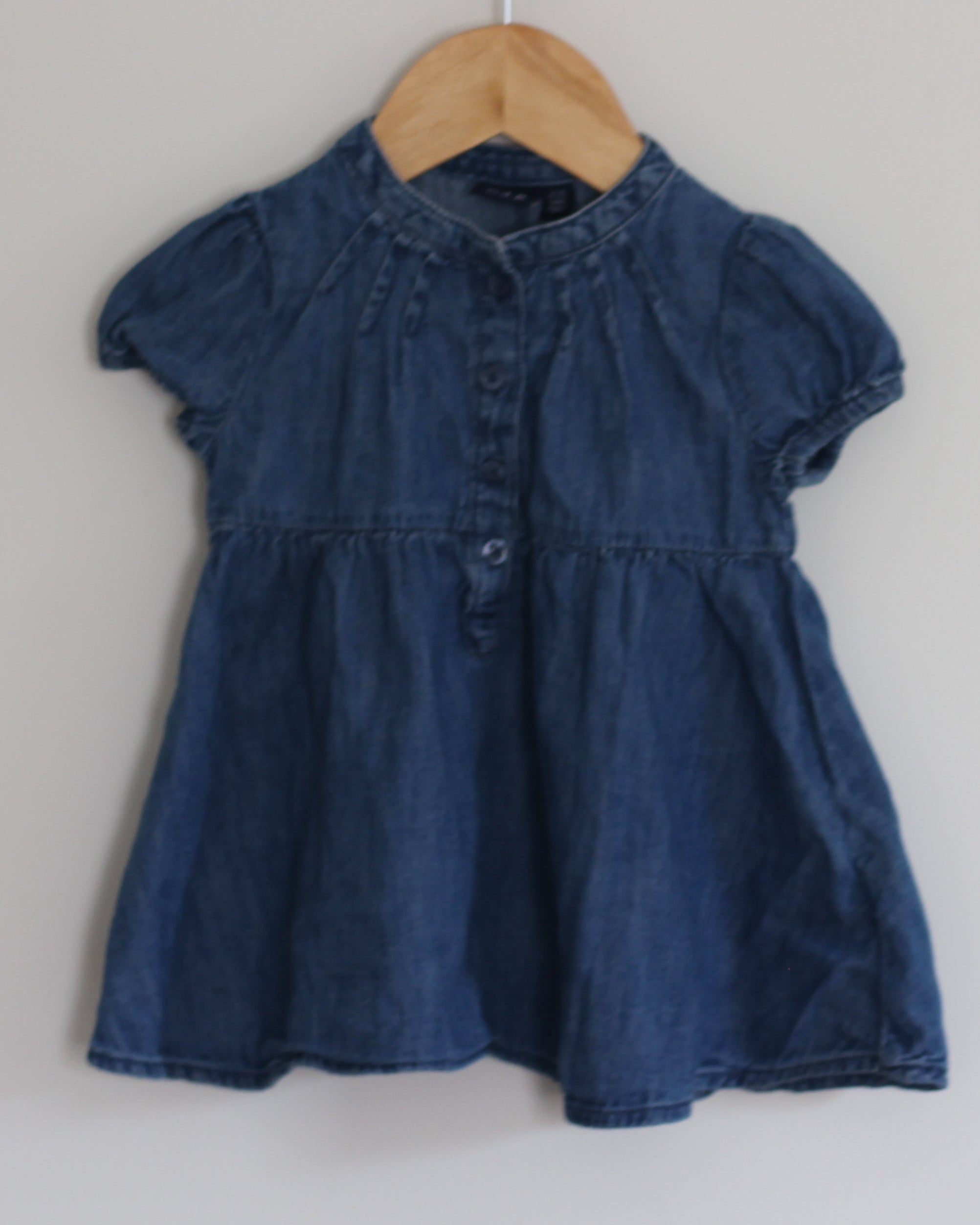 A short-sleeve blue denim dress hanging on a wooden hanger from a wall hook against a plain white wall.