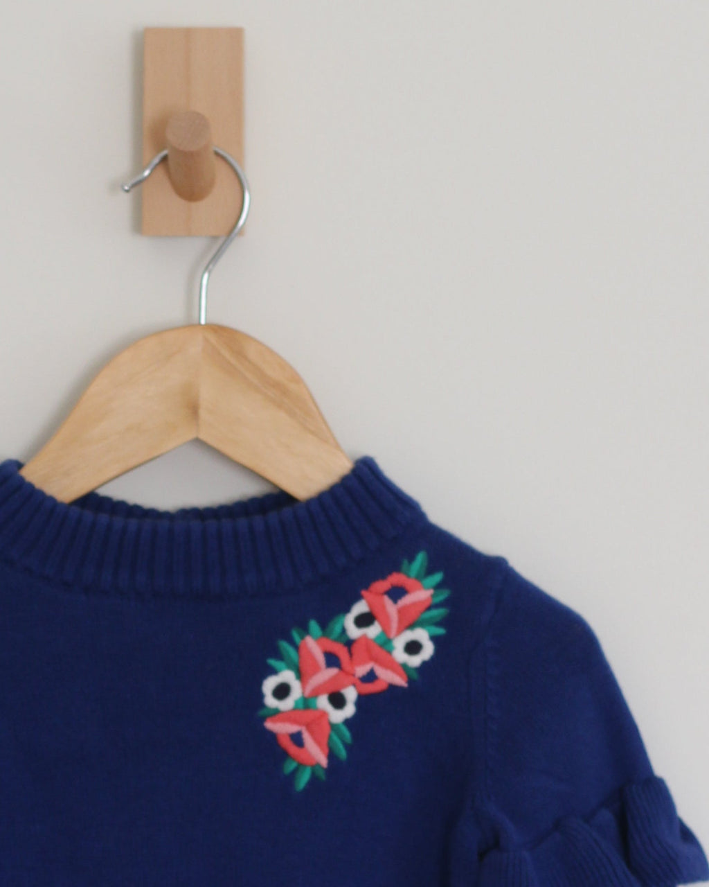 A navy blue knit sweater with ruffled sleeves and colorful floral embroidery on the chest, hanging on a wooden hanger from a wall hook.