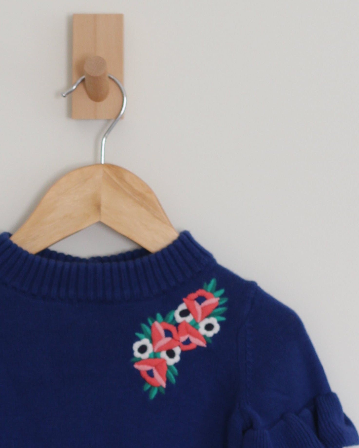 A navy blue knit sweater with ruffled sleeves and colorful floral embroidery on the chest, hanging on a wooden hanger from a wall hook.
