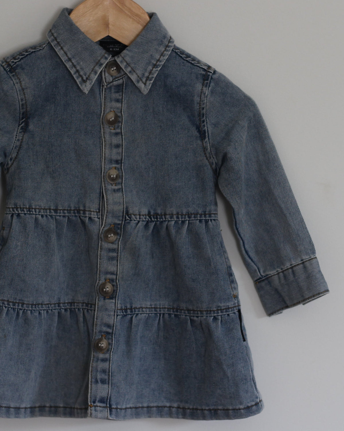 A toddler's blue denim shirt dress with long sleeves and a button-up front, hanging on a wooden hanger from a wall hook against a plain light gray wall.