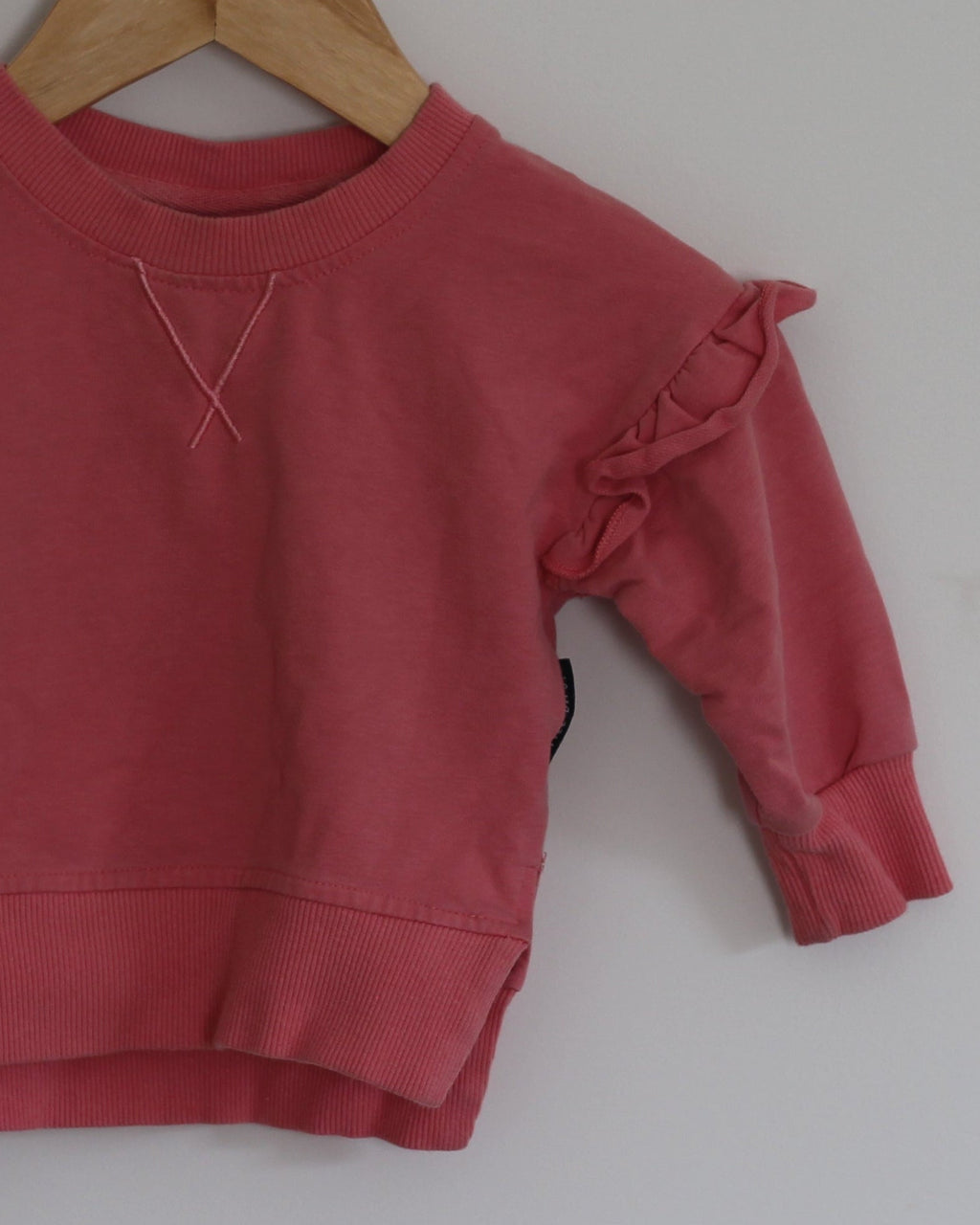 A dusty rose pink long-sleeve sweatshirt with ruffled shoulder details, hanging on a wooden hanger from a small wooden peg on a plain light gray wall.