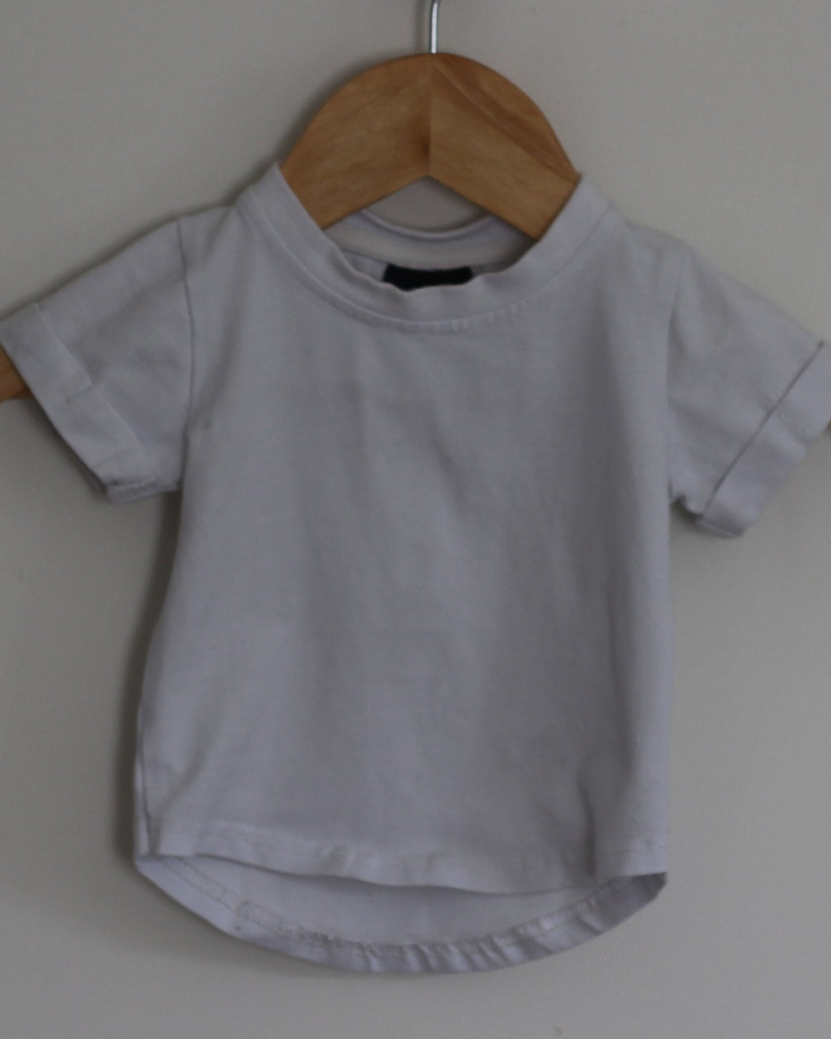 White short-sleeve t-shirt hanging on a wooden hanger from a small wooden peg on a plain off-white wall.